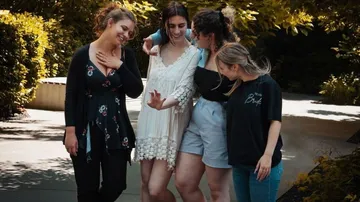 Four women stand together, laugh and interact outdoors.