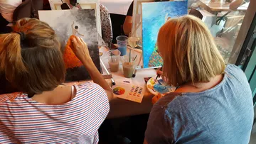 Two women paint canvases together in a café.