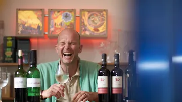 Laughing man holding a wine glass in front of wine bottles.