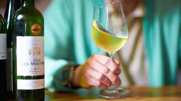 A glass of wine is held by one hand, surrounded by bottles.
