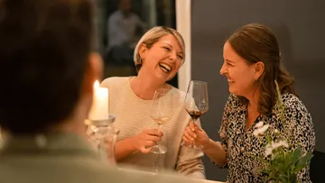 Two women laugh and toast with glasses, cozy table atmosphere.