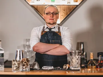 Barkeeper steht mit verschränkten Armen hinter der Bar.