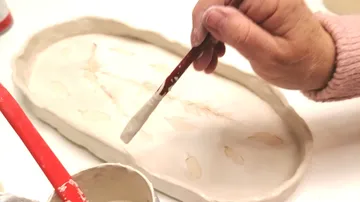Eine Hand malt mit einem Pinsel auf keramischer Oberfläche.