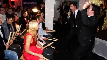 Menschen schlagen auf Eimer, während Sänger performt. Feierliche Atmosphäre.