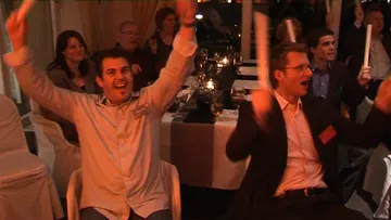 Zwei Männer jubeln mit weißen Stäben in einem festlichen Raum.