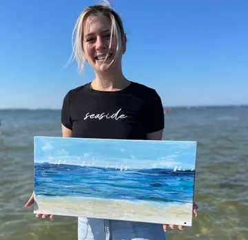 Woman holding a painting of a beach in the sun.