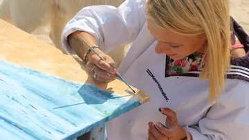 Frau malt konzentriert auf einer Holzfläche am Strand.