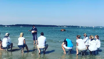 Gruppe sitzt am Strand, während jemand ihnen etwas erklärt.
