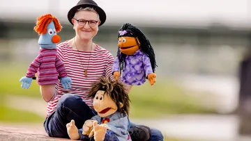 Puppenspieler sitzt mit vier bunten Puppen im Freien.