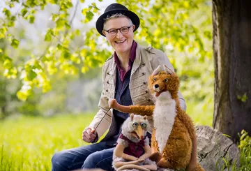 Person sitzt lächelnd mit zwei Handpuppen im Freien.