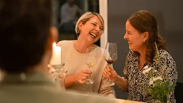 Two women laugh and toast with wine glasses, cozy atmosphere.