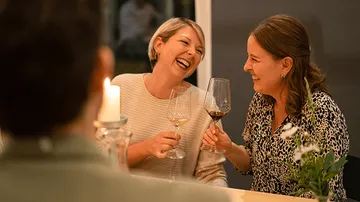 Two women laugh and toast with wine glasses, by candlelight.