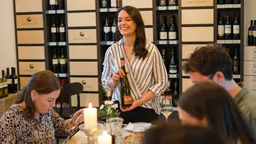 Woman presenting wine bottle, guests at table, relaxed atmosphere.