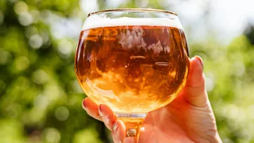 Ein Glas mit goldenem Getränk wird gehalten, im Freien.