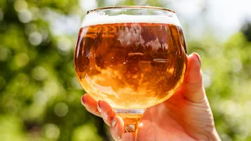 Ein Glas mit goldenem Getränk wird gehalten, im Freien.