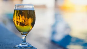 A glass of beer stands on a rocky surface by the water.