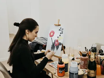 A woman paints flowers on canvas surrounded by colors.