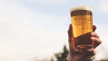 A glass of beer is held up, sky in the background.