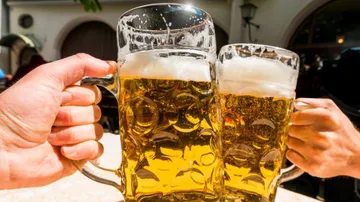 Two beer mugs toast, surrounded by people outdoors.