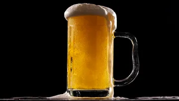 A beer mug overflows with frothy beer against a black background.