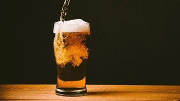 A glass of beer is filled in front of a dark wall.