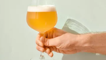 Ein Hand hält ein Glas Bier vor einer hellen Wand.