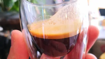 A glass of espresso is held by one hand.