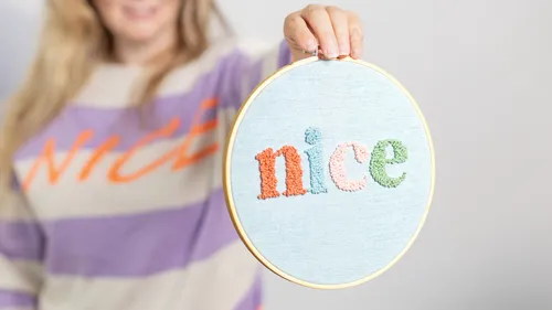 Gestickter Rahmen mit dem Wort „nice“ wird gehalten.