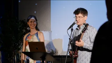 Musicians play live; a man sings, a woman smiles.