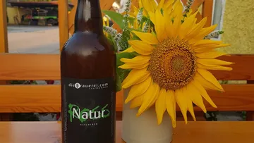 Beer bottle stands next to sunflower on wooden outdoor table.