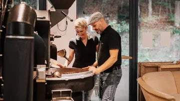 Kaffeeröster zeigt Bohnen, Mitarbeiter beobachten in gemütlicher Umgebung.