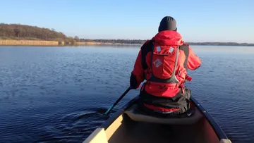Person paddelt im Kanu auf ruhigem Wasser unter blauem Himmel.