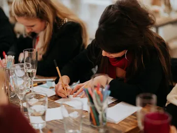 Women draw at tables with drinks and pens.