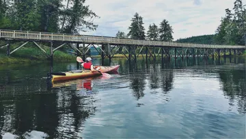 Kajakfahrer paddelt ruhig unter einer Holzbrücke in der Natur.