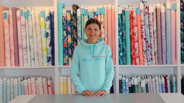 Young woman stands in front of colorful rolls of fabric in a shop.