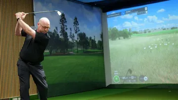 Mann schwingt Golfclub in einer Indoor-Umgebung mit Bildschirm.