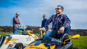 Three men pose with toy cars in a landscape.