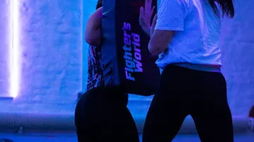 Person hält Schlagpolster, andere trainiert im Fitnessstudio.