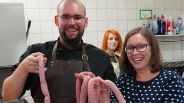 Two people are holding sausages in a kitchen setting, smiling.