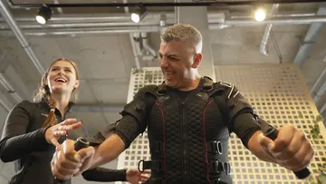 Mann trainiert mit EMS-Anzug, Frau hilft in Fitnessstudio.