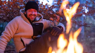 Mann sitzt lächelnd neben einem Feuer im Wald.