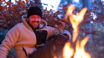 Man sitting next to a campfire in the forest, smiling.