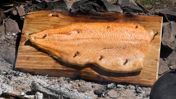 Salmon fillet is cooked over fire on a wooden board.