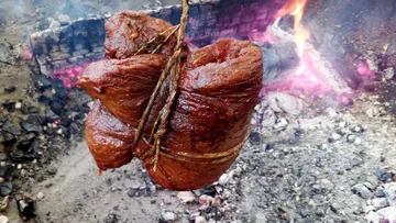 A piece of meat hangs over glowing charcoal in the open.