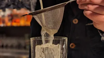 Ein Sieb filtert Flüssigkeit in ein Glas mit Eis.