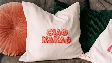 Ein Kissen mit roten Buchstaben „CIAO KAKAO“ liegt auf einer Couch.