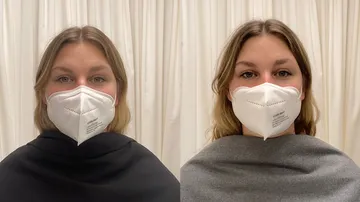 Two women wear white masks and stand in front of a curtain.