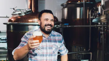 A man is smiling and holding a beer in a brewery.