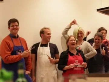 Gruppe kocht zusammen, lacht mit Schürzen und Utensilien.