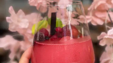 Glas mit pinkem Getränk und Früchten vor Blumenarrangement.